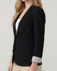 Woman wearing a black blazer over a white shirt with beige pants on a light gray background