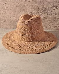 A light tan woven straw hat with a textured geometric pattern, displayed on a neutral concrete surface against a textured concrete wall.