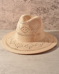 A light beige crocheted wide-brim hat with a structured crown and geometric openwork patterns, displayed on a textured concrete surface against a neutral background.