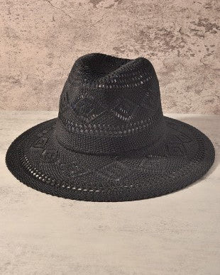 A black woven fedora hat with an intricate openwork pattern, displayed against a textured beige and gray background.