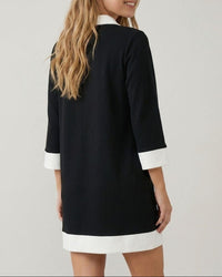 A woman wearing a black three-quarter sleeve tunic dress with white contrast trim at the collar, cuffs, and hem, shown from a back-three-quarter angle against a light gray studio background.