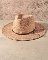A woven straw fedora hat in a light tan color with a dark brown band, displayed on a textured beige surface against a rustic, distressed beige wall.