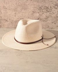 An off-white woven straw cowboy hat with a dark brown band and matching chin strap, displayed on a textured light gray surface against a matching gray background.