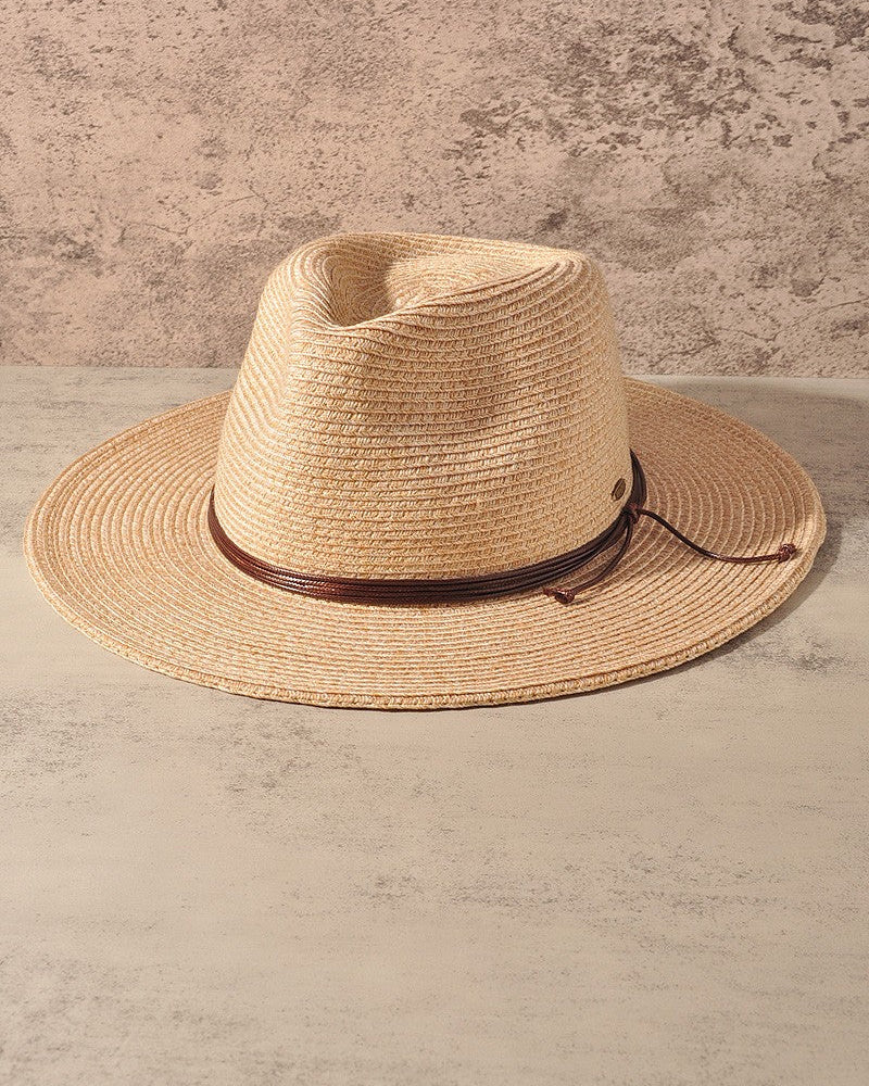 A natural straw fedora hat with a dark brown leather band, displayed on a textured beige surface against a matching textured wall.
