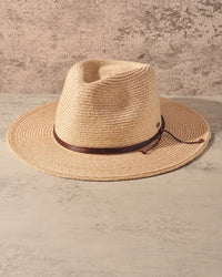 A natural straw fedora hat with a dark brown leather band, displayed on a textured beige surface against a matching textured wall.